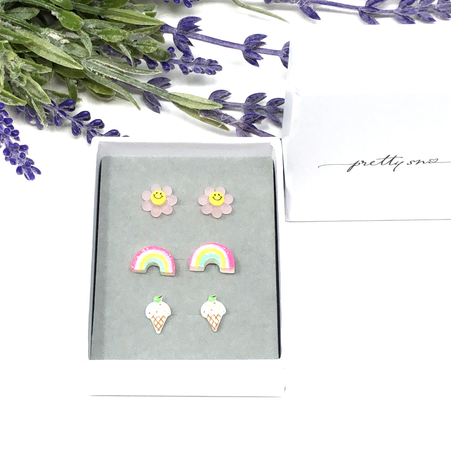 Three pairs of earrings displayed in a box, featuring a pink smiley face, a multicolored rainbow, and a vanilla ice cream cone design.