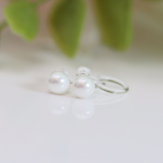 A pair of invisible clip-on pearl earrings displayed on a white surface with a green leaf in the background.