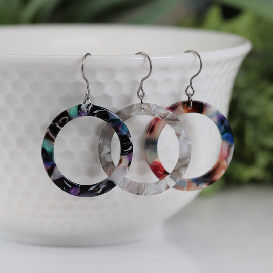 A pair of multicolor acrylic circle hoop dangle earrings displayed over a white bowl with green leaves in the background.