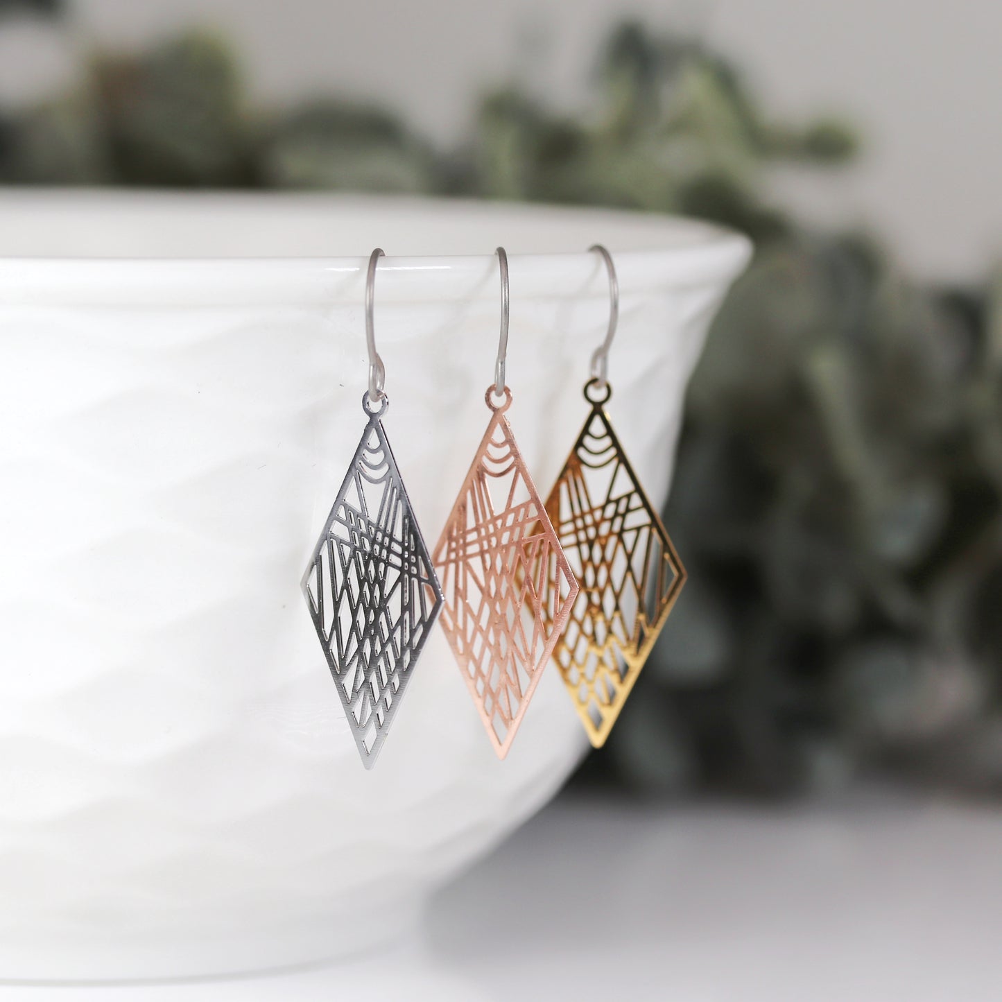 Three geometric dainty rhombus dangle earrings in silver, rose gold and gold color, displayed over a white textured surface.