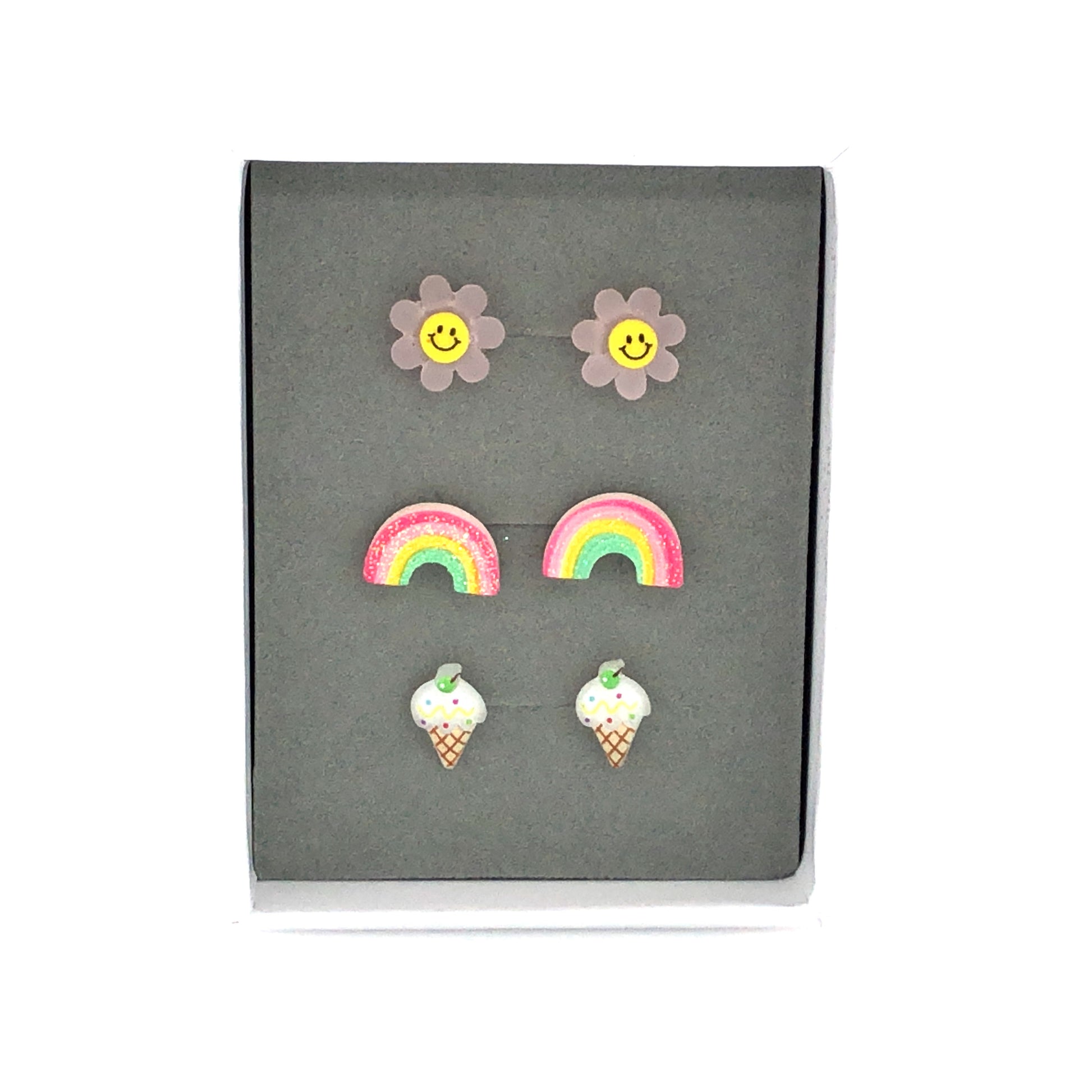 Three pairs of earrings displayed in a box, featuring a pink smiley face, a multicolored rainbow, and a vanilla ice cream cone design.