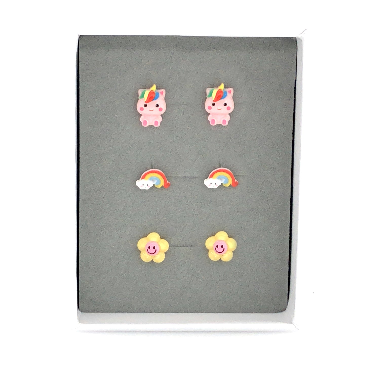 A set of three pairs of stud earrings featuring a pink rainbow unicorn, a dainty rainbow with cloud, and a yellow and pink smiley face flower, presented in a grey box.