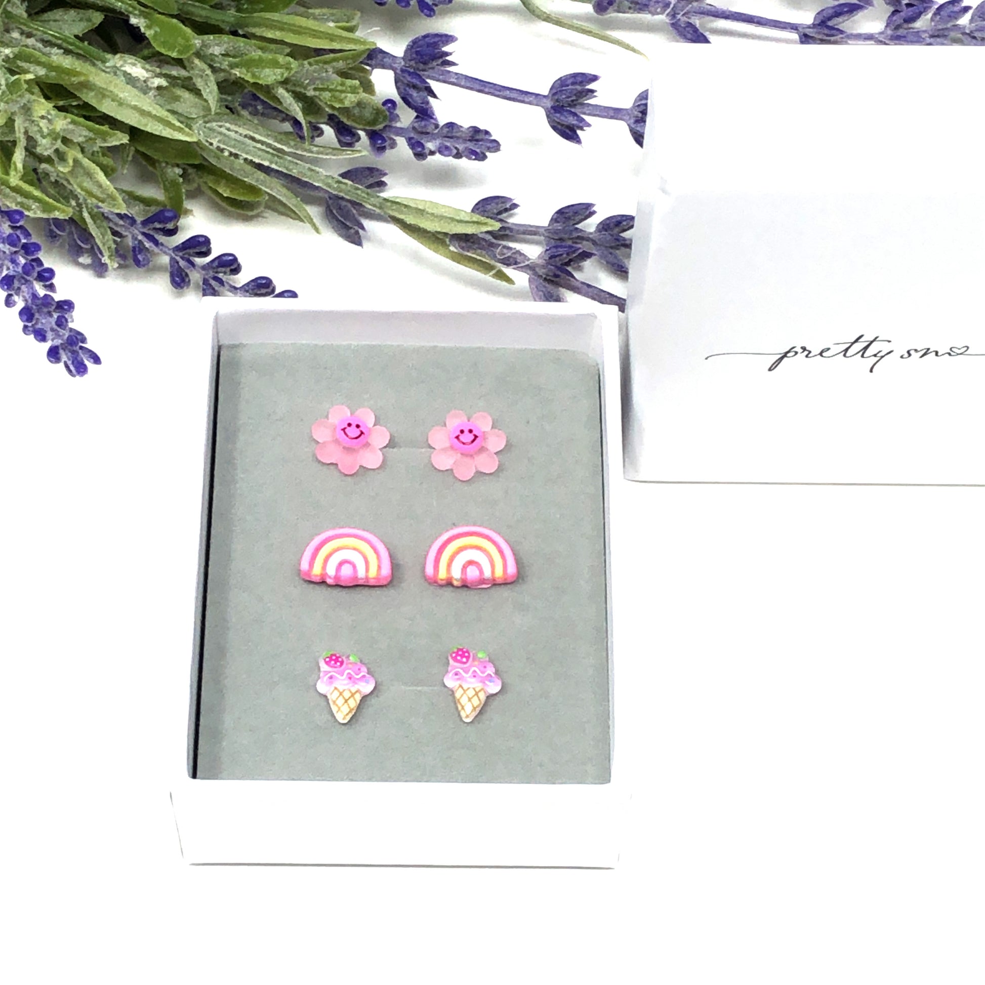Three pairs of earrings featuring a dark pink smiley face daisy, a pink rainbow, and a purple ice cream cone design, presented in a box 
