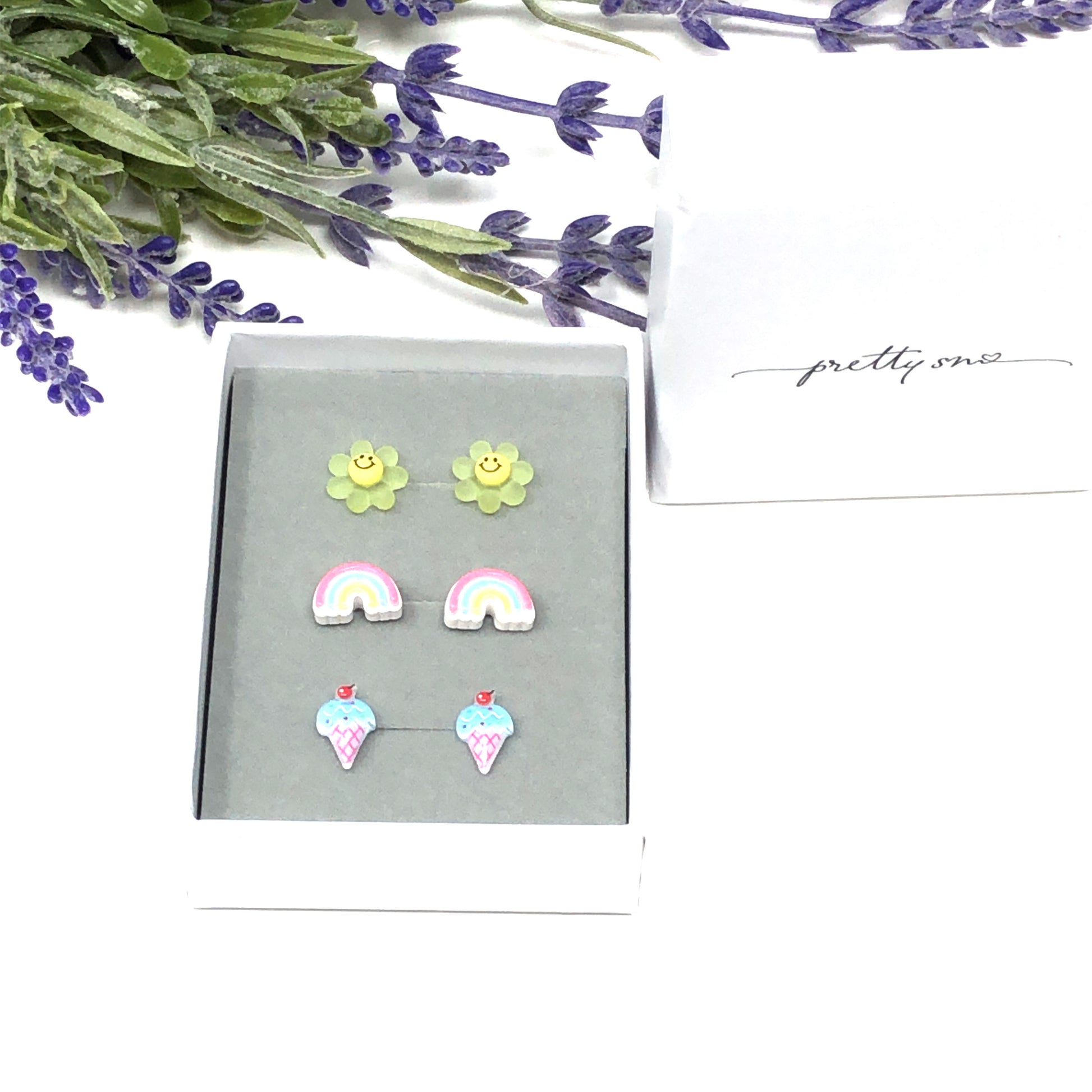 Three pairs of earrings displayed in a box, featuring a yellow smiley face, a pastel rainbow, and a blue ice cream cone design.