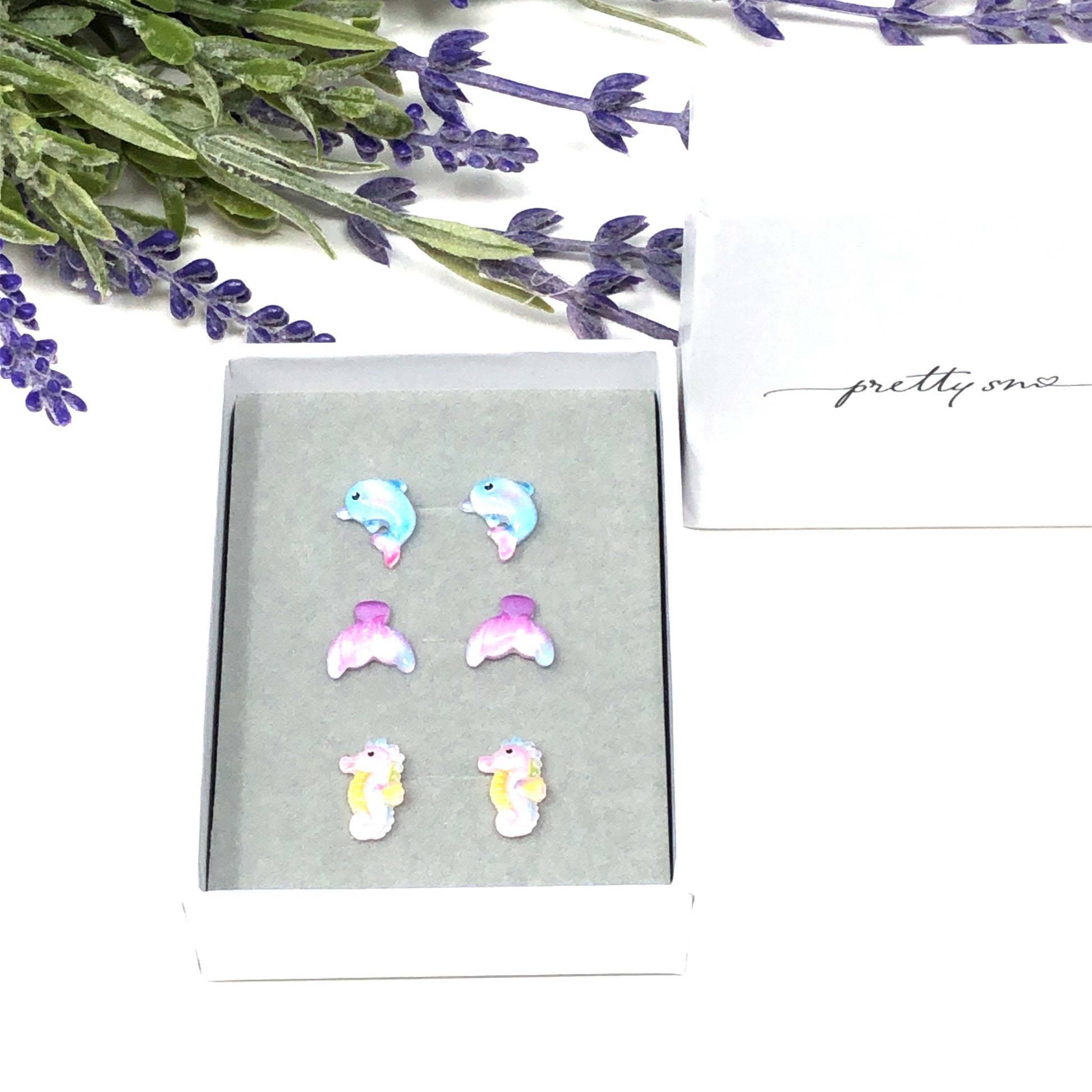 A set of three pairs of stud earrings featuring resin animals: a dolphin, a mermaid tail, and a seahorse, presented in a gift box.