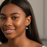 Close-up of an ear wearing a pink heart-shaped earring with a blue accent, with blurred flowers in the background.