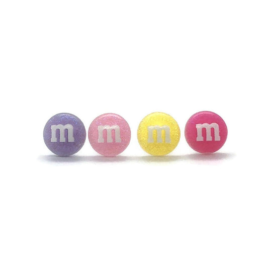 A set of four pastel-colored M&M candy stud earrings arranged in a line, with colors including purple, pink, yellow, and pink again, all with the iconic M&M 'M' logo on them.