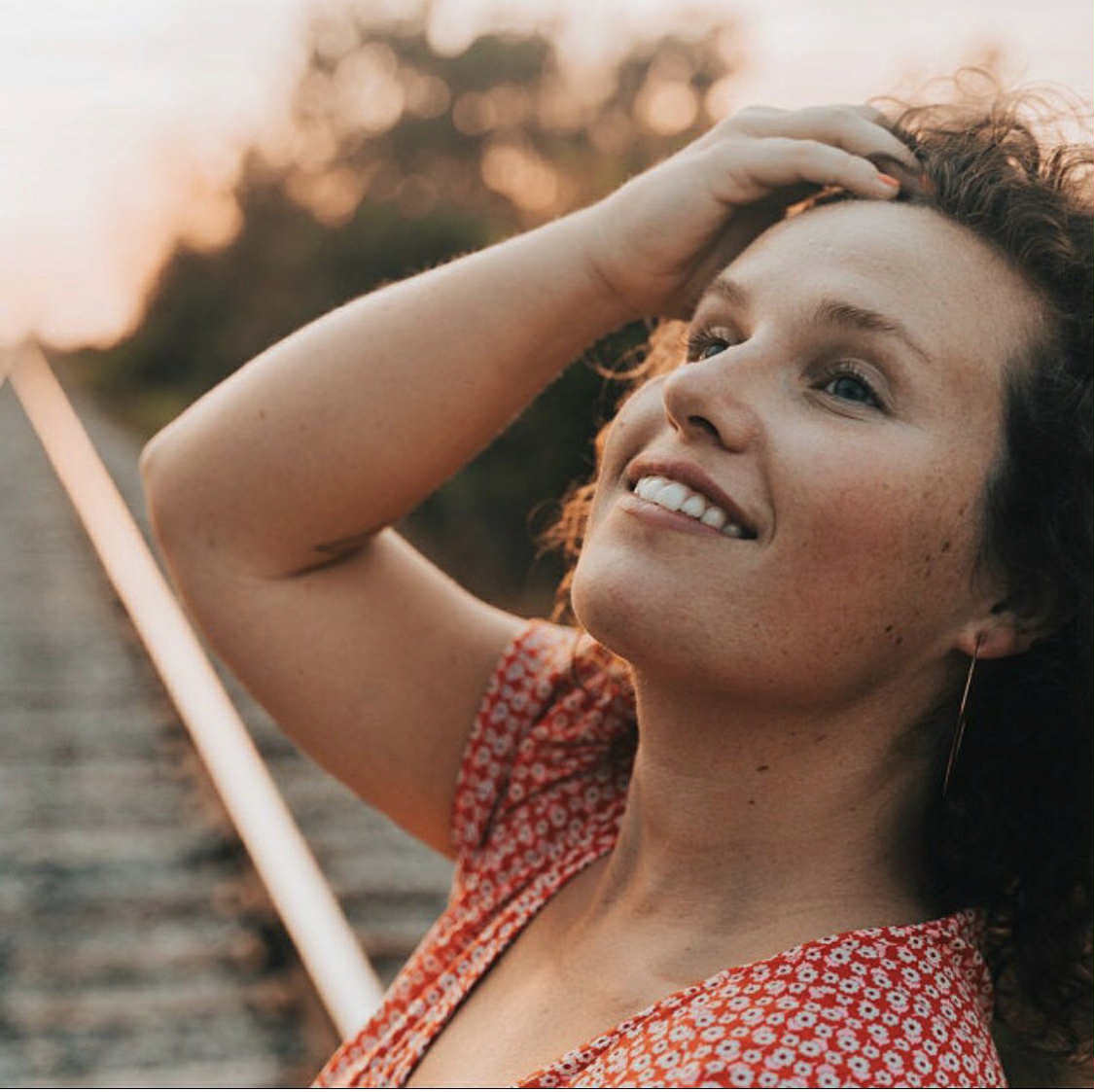 Woman in a red floral dress smiling outdoors with a blurred background wearing large wire hoops 