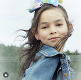 Young girl with a denim jacket and colorful hair bow outdoors with pink glitter unicorn earrings