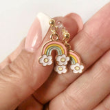 Rainbow and flower earrings held between fingers against a light background