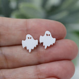 White ghost-shaped earrings held between fingers with a blurred green background