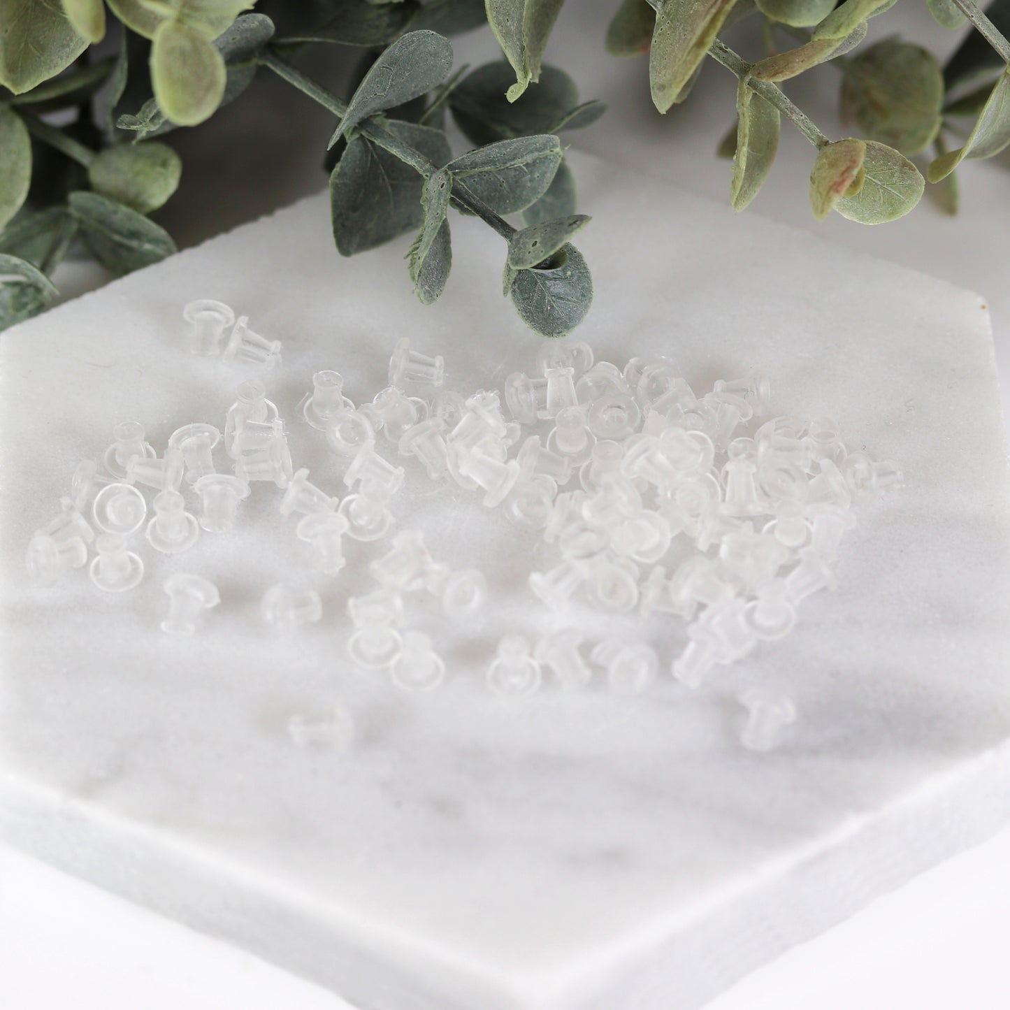 A collection of clear rubber ear nut backs for earrings displayed on a white surface, with green leaves in the background.