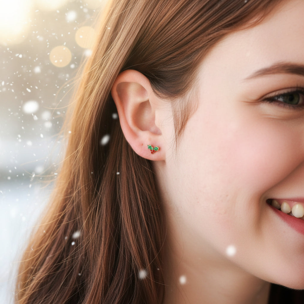 A pair of dainty holly stud earrings with a gold PVD coating and red, green, on ear