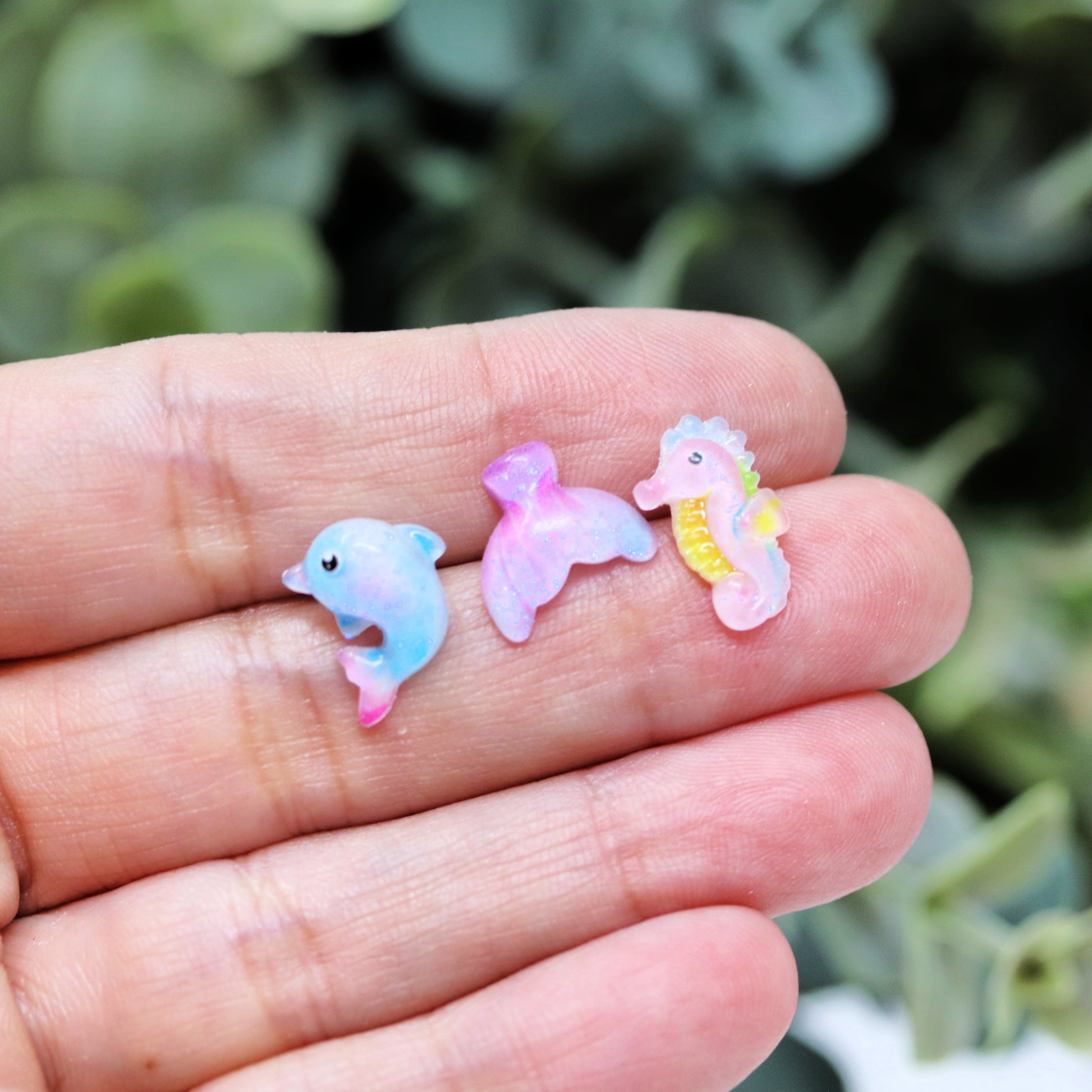A set of three pairs of stud earrings featuring resin animals: a dolphin, a mermaid tail, and a seahorse, presented in a hand