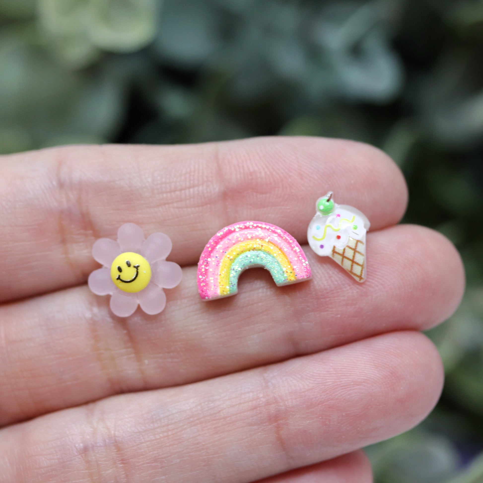 Three pairs of earrings displayed in a box, featuring a pink smiley face, a multicolored rainbow, and a vanilla ice cream cone design in hand