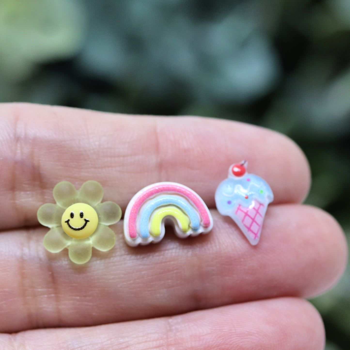 Three pairs of earrings displayed in a box, featuring a yellow smiley face, a pastel rainbow, and a blue ice cream cone design.