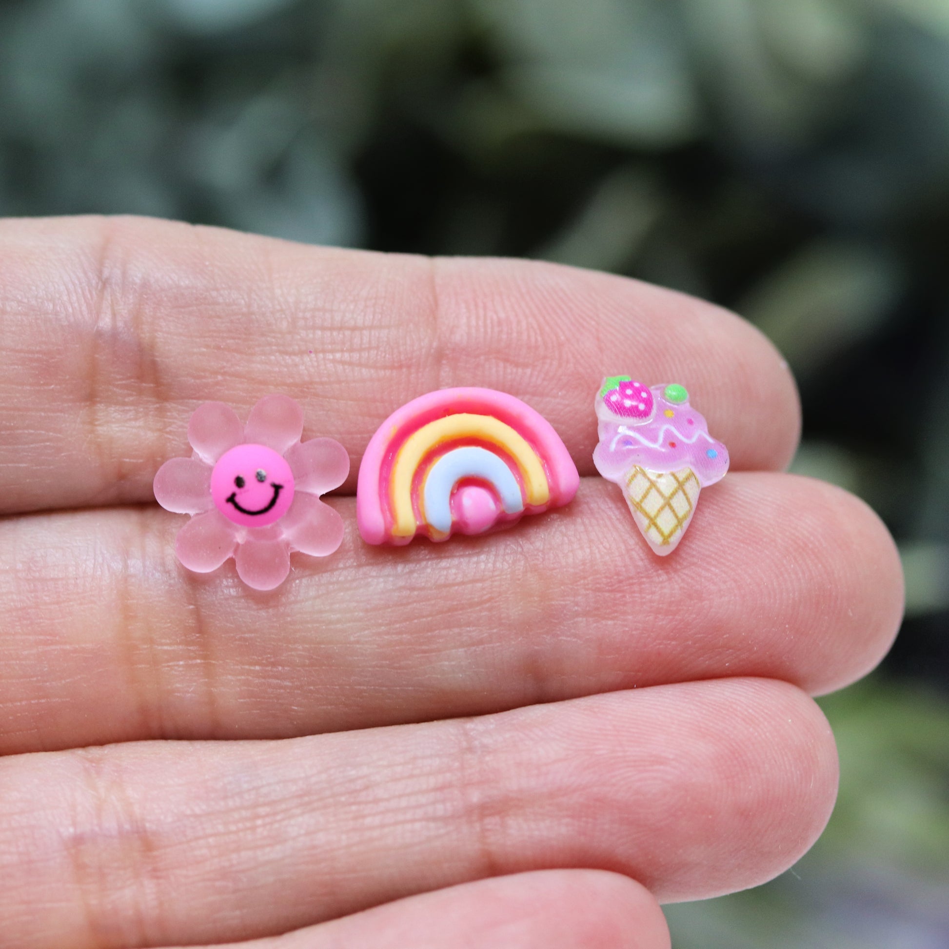 Three pairs of earrings featuring a dark pink smiley face daisy, a pink rainbow, and a purple ice cream cone design, 