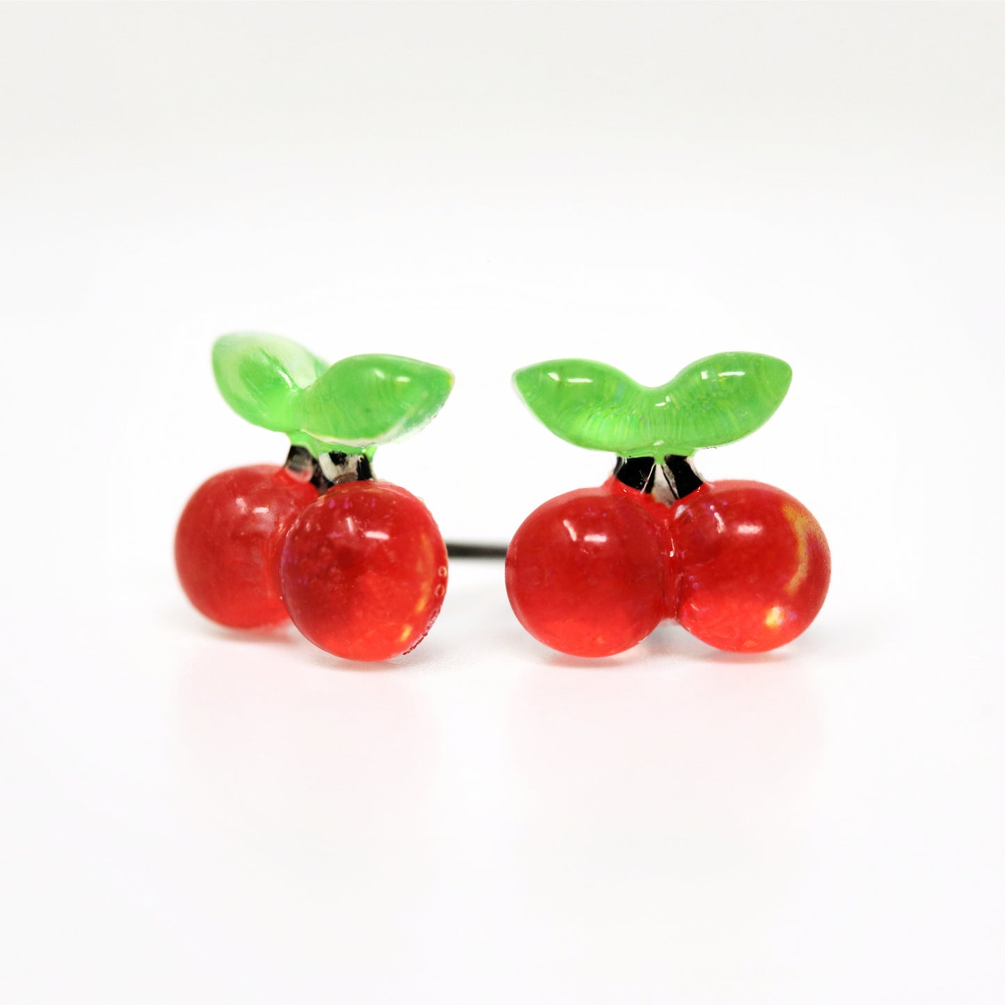 A pair of red cherry-shaped stud earrings with green leaves on top, attached to white posts.