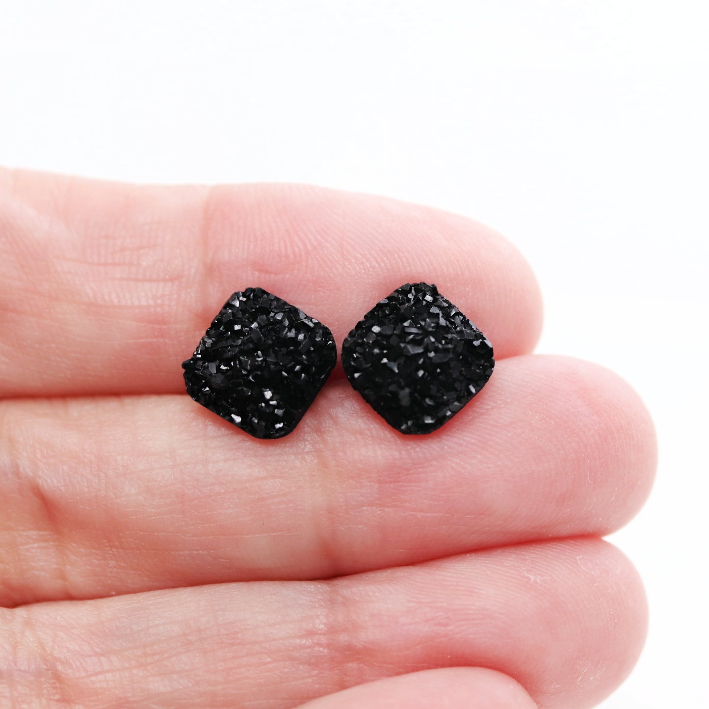 A pair of black square druzy stud earrings with rounded corners on a white background