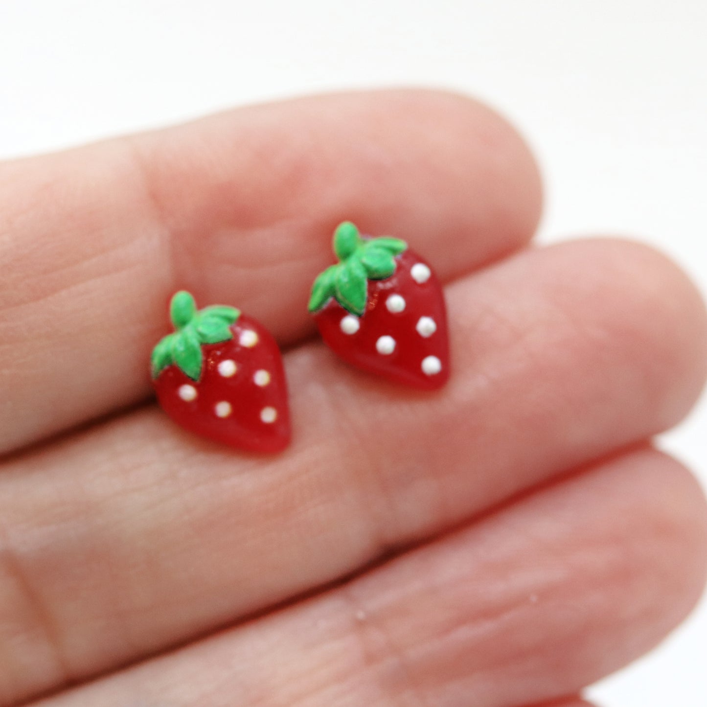 A pair of red strawberry-shaped stud earrings with green accents and white dots on a white background.