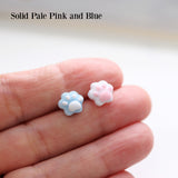 Two small paw-shaped earrings, one pink and one blue, held between fingers against a neutral background.