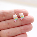 Small mermaid cat-shaped earrings with rainbow tails held between fingers on a white background