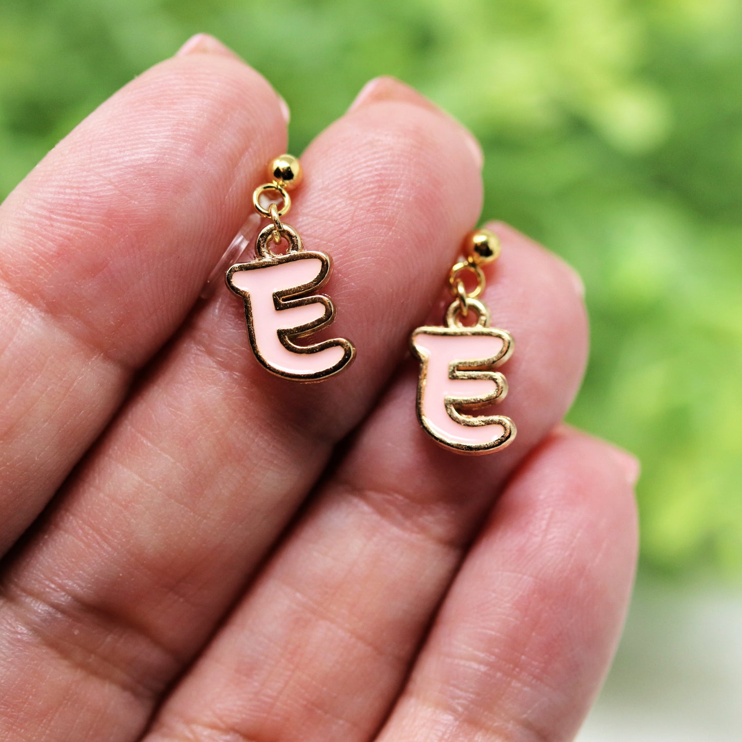 Gold earrings with pink 'E' charms held between fingers against a blurred green background