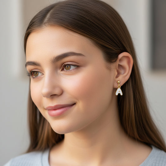 Gold earrings with 'A' charms held between fingers against a blurred green background