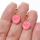 Pink smiley face earrings with gold accents held between fingers on a white background