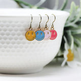 Set of smiley face earrings in yellow, blue, pink, and red hanging on a white teacup with a plant in the background.