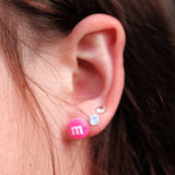Close-up of an ear wearing two earrings: a pink 'm&M' logo earring and a silver rhinestone earring.