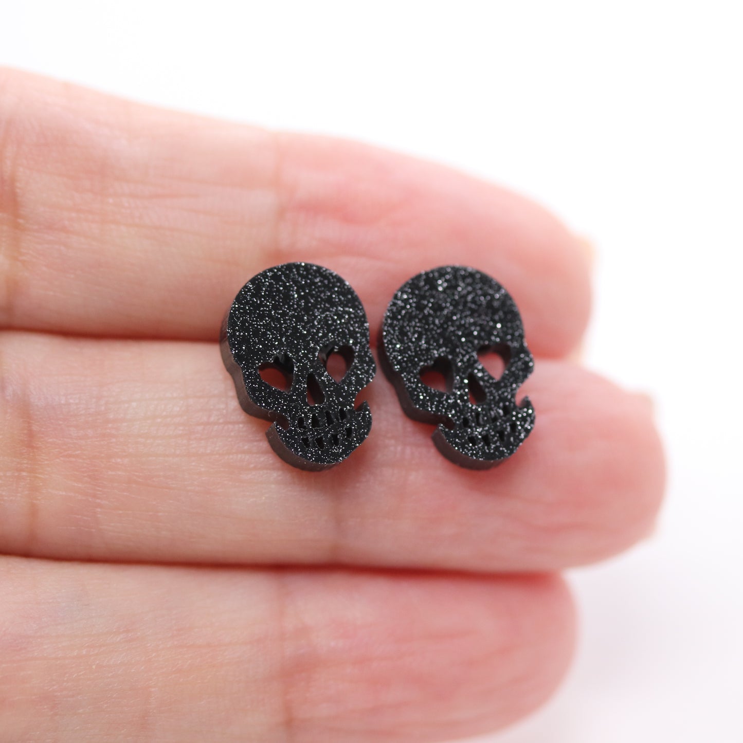Black glittery skull-shaped earrings held between fingers on a white background