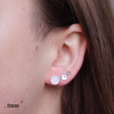 Close-up of an ear wearing small white opalescent resin earrings with a visible size measurement.