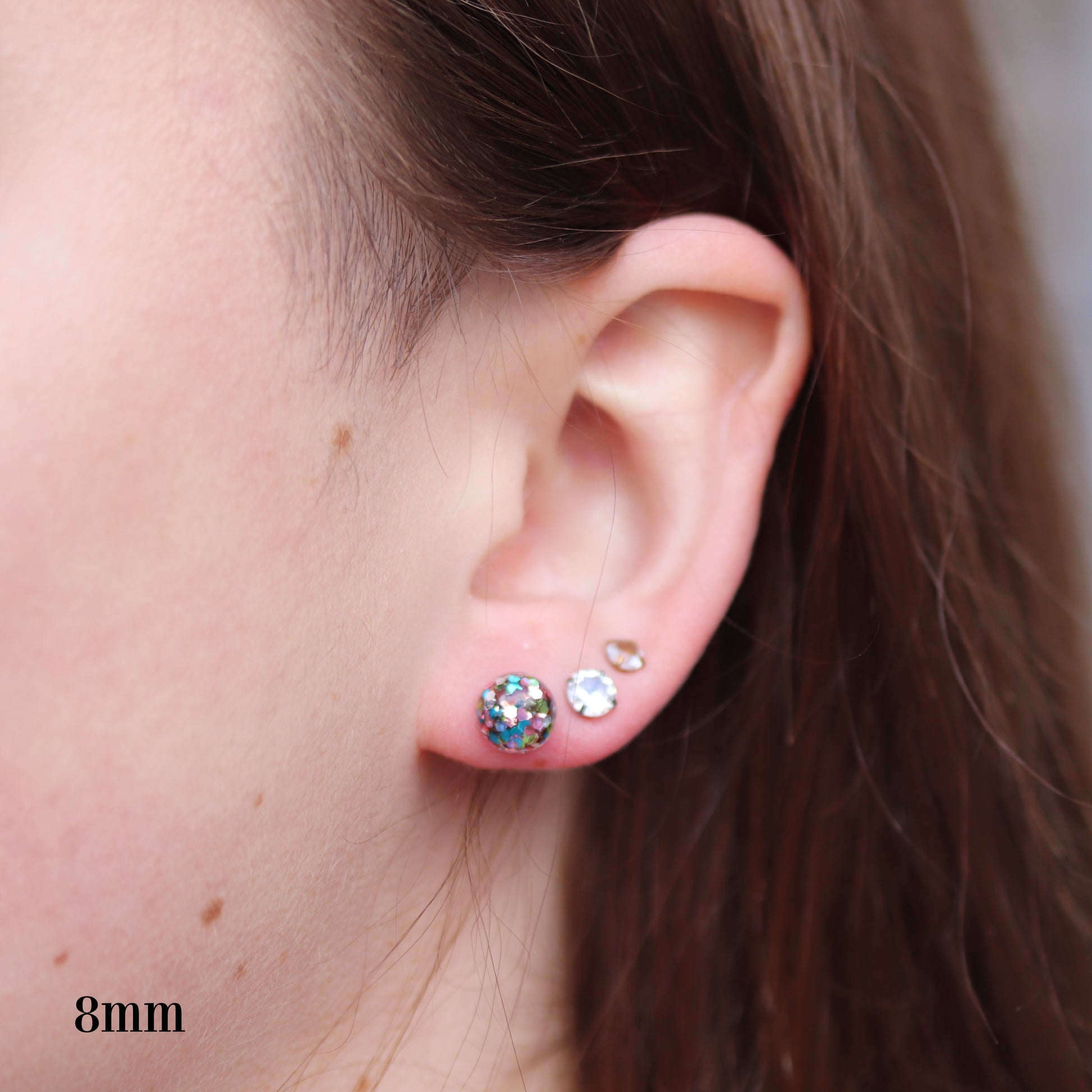 Close-up of an ear wearing small glitter dome earrings with colorful confetti flakes.