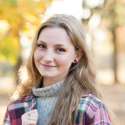 Woman with long blonde hair wearing 8mm burgundy faux druzy studs and a plaid shirt outdoors with blurred autumn trees in the background