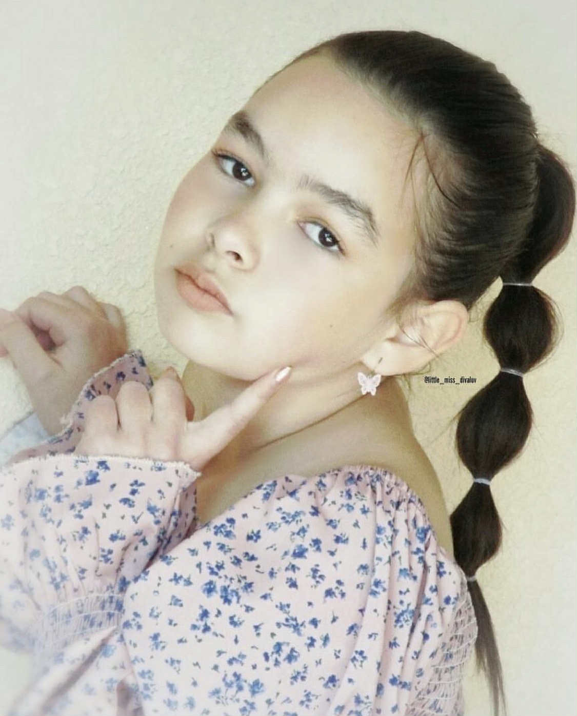Young girl with braided hair wearing a floral dress and a butterfly dangle earring against a plain background