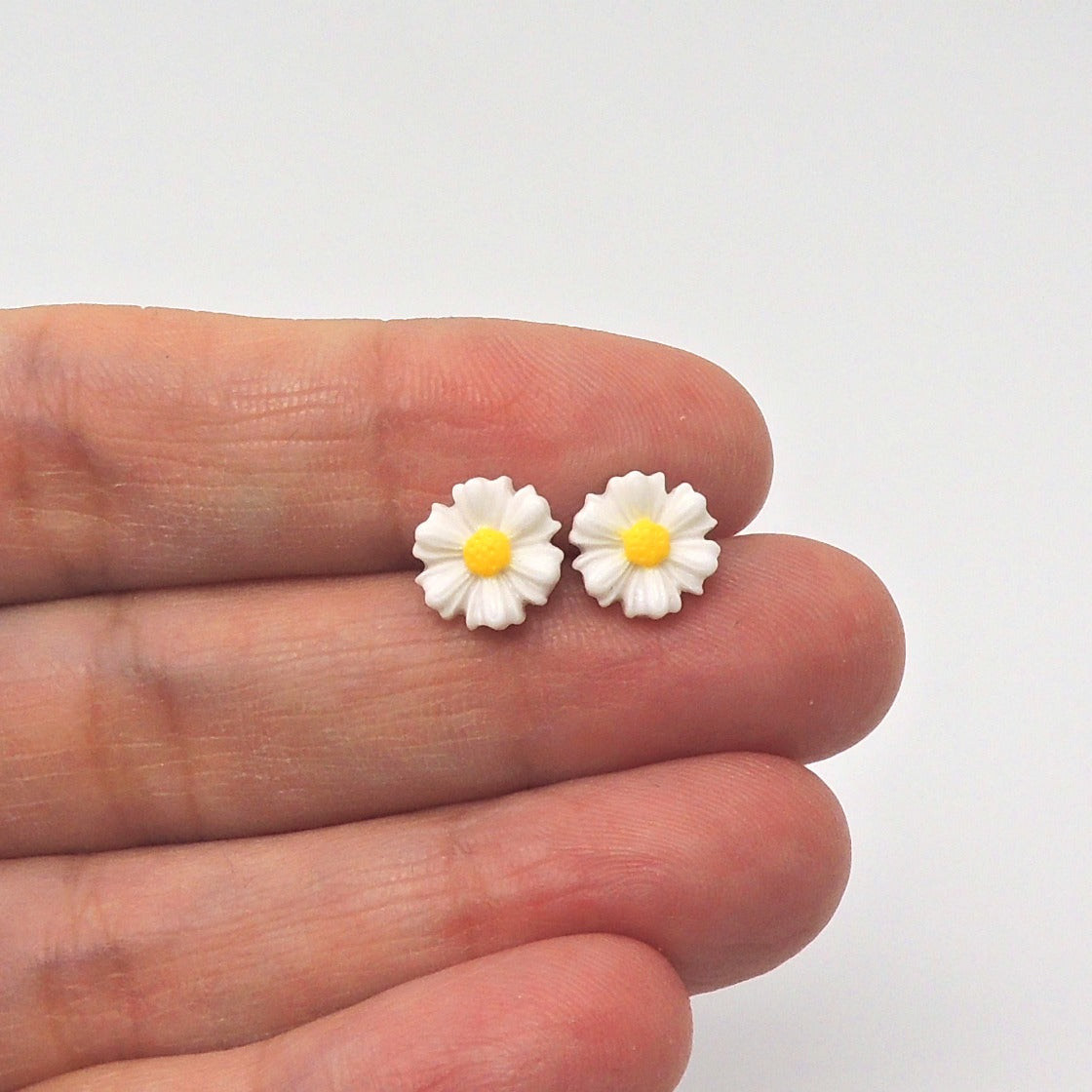 A pair of daisy stud earrings in white and yellow, held between fingers against a white background.
