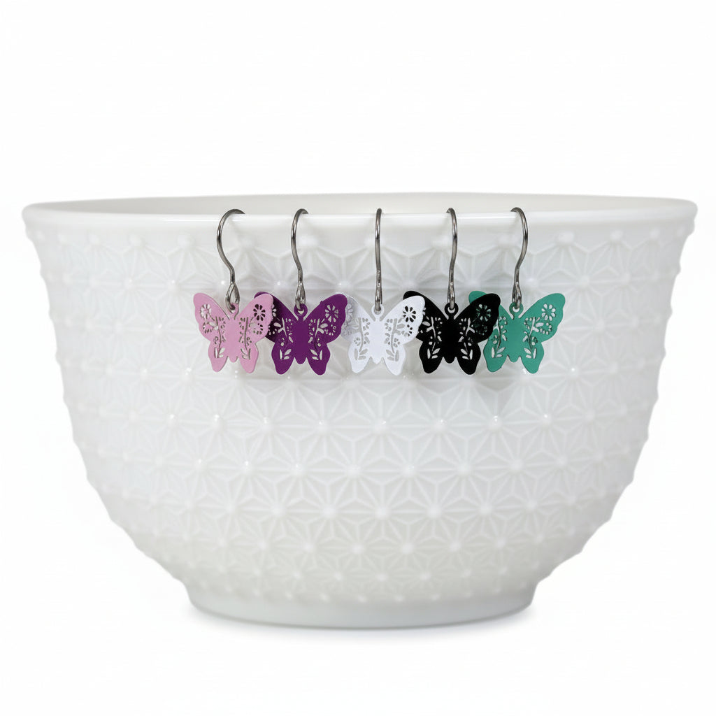 A set of dangle earrings with butterfly designs, displayed in a white bowl with a geometric pattern. The earrings come in various colors including black, white, pink, purple, and green.