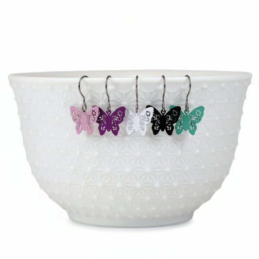 A set of dangle earrings with butterfly designs, displayed in a white bowl with a geometric pattern. The earrings come in various colors including black, white, pink, purple, and green.