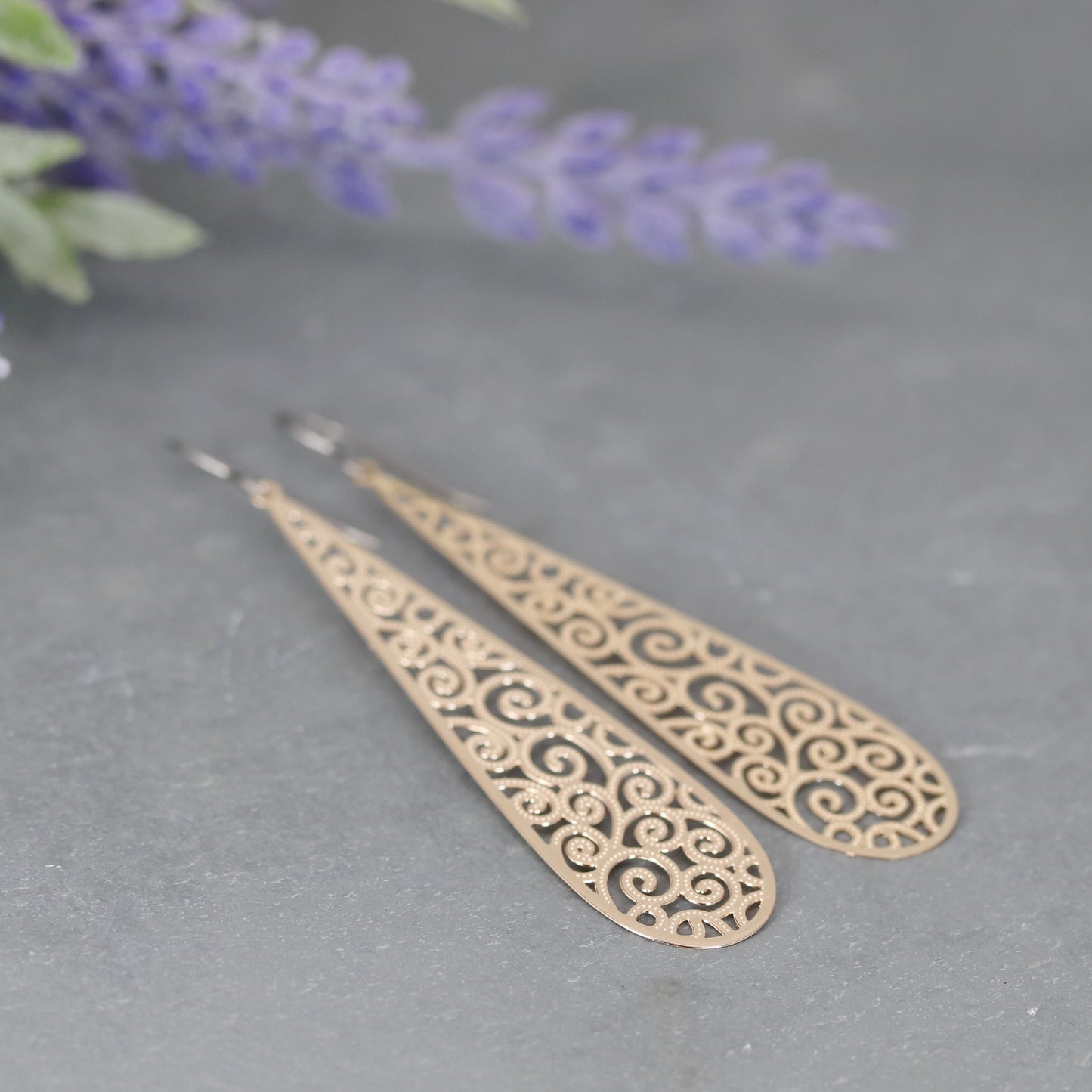 Long teardrop dangle earrings with a filigree design, made of stainless steel, displayed on a textured surface with purple flowers in the background.