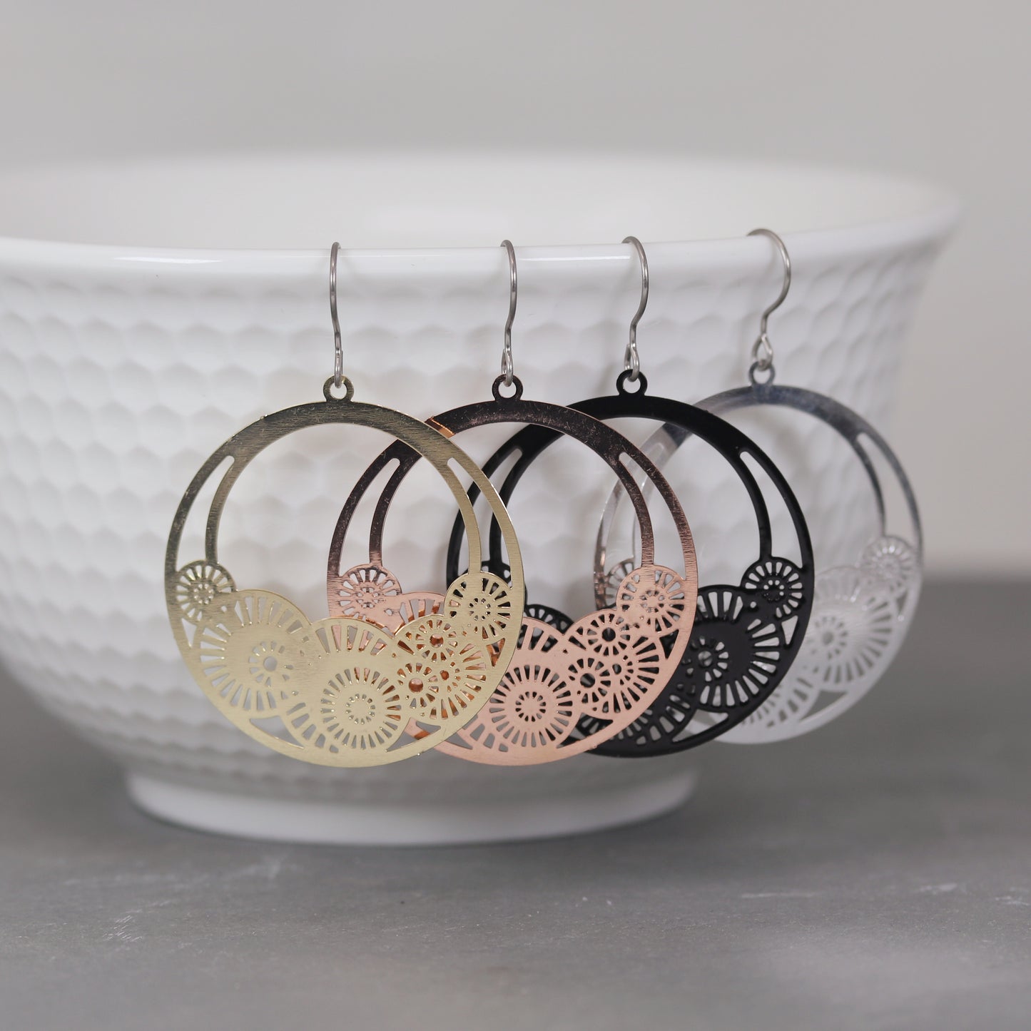 A pair of dangle earrings with laser cut out circles, displayed above a white bowl. The earrings are in gold and silver colors, with rose gold and black variations.