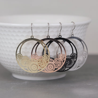 A pair of dangle earrings with laser cut out circles, displayed above a white bowl. The earrings are in gold and silver colors, with rose gold and black variations.