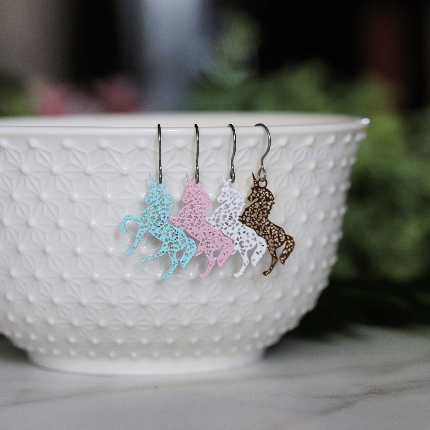 A set of dangle earrings in the shape of unicorns, displayed in a white bowl, with variations in colors like pale blue, white, pale pink, and gold.