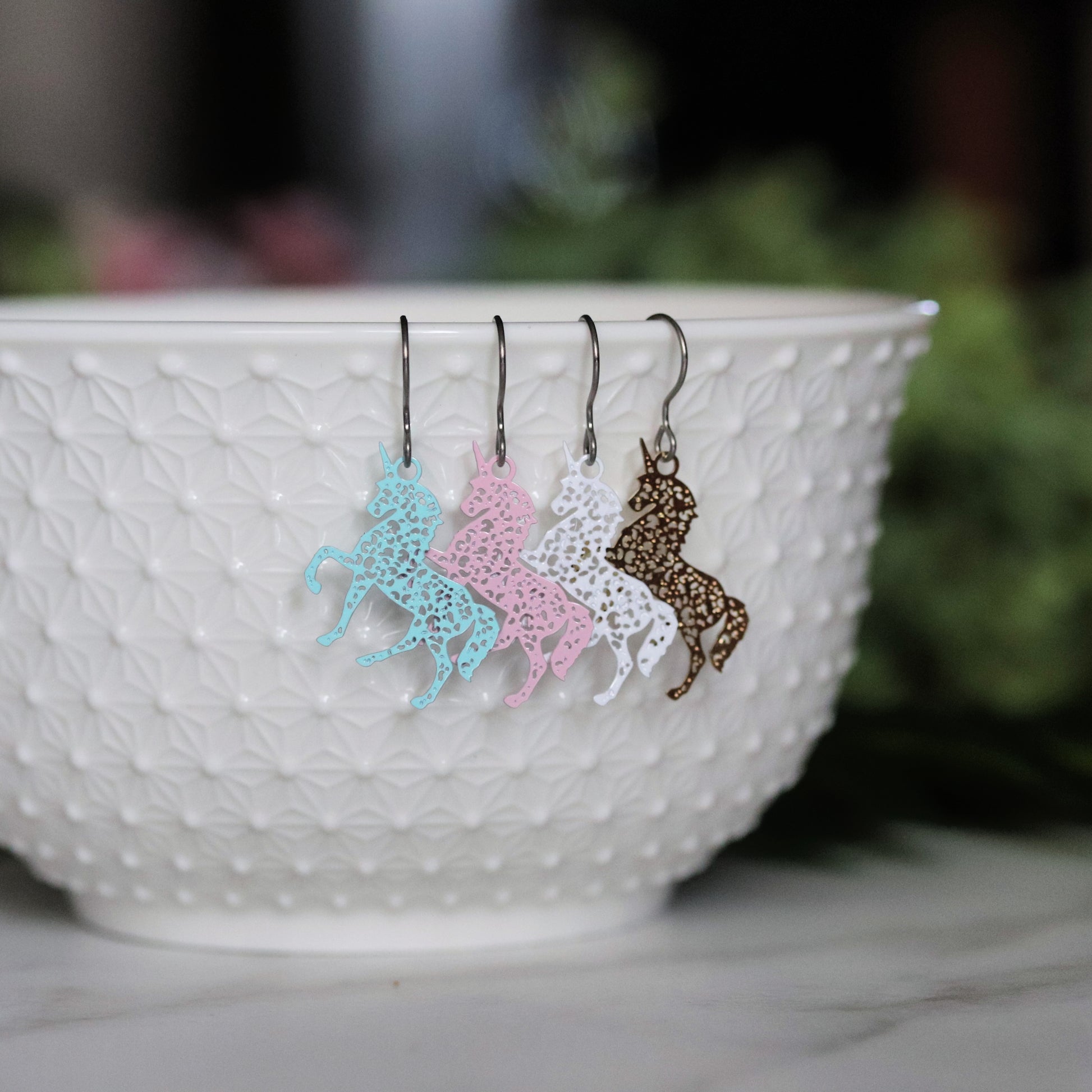 A set of dangle earrings in the shape of unicorns, displayed in a white bowl, with variations in colors like pale blue, white, pale pink, and gold.