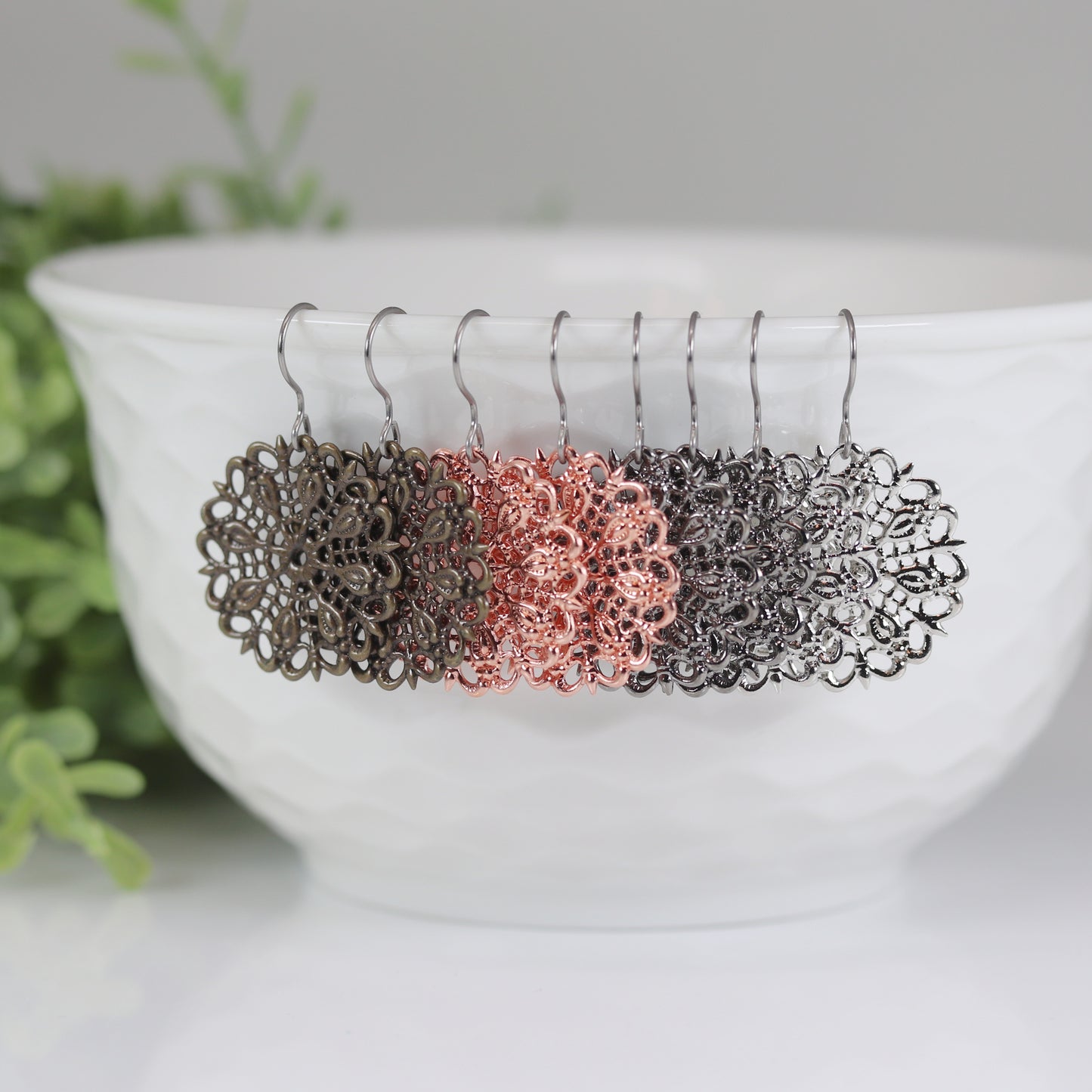 A set of dangle earrings in silver and bronze colors displayed on a white earthenware bowl, with a green plant in the background.