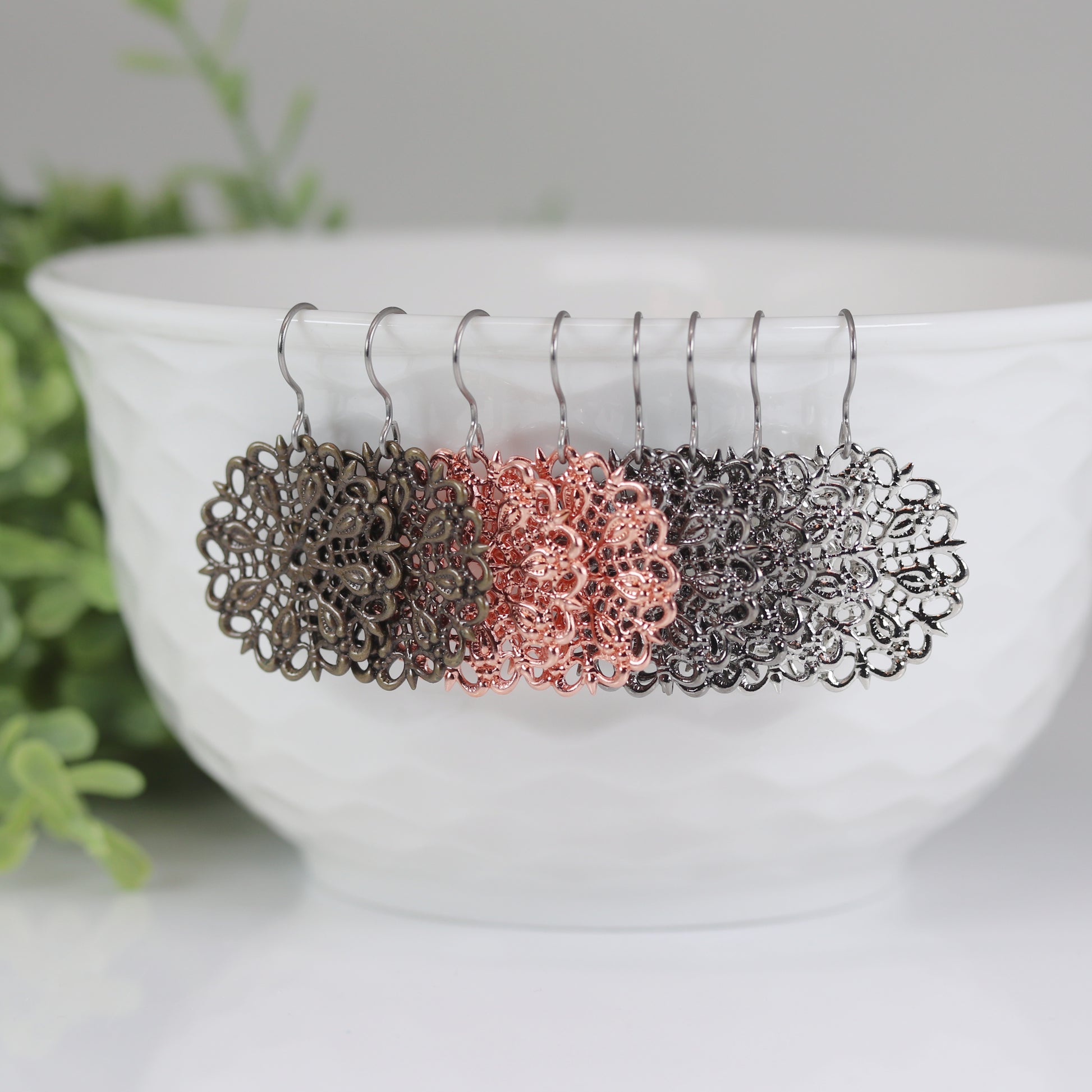 A set of dangle earrings in silver and bronze colors displayed on a white earthenware bowl, with a green plant in the background.