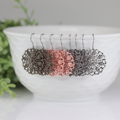 A set of dangle earrings in silver and bronze colors displayed on a white earthenware bowl, with a green plant in the background.
