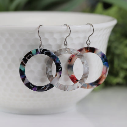 A pair of multicolor acrylic circle hoop dangle earrings displayed over a white bowl with green leaves in the background.