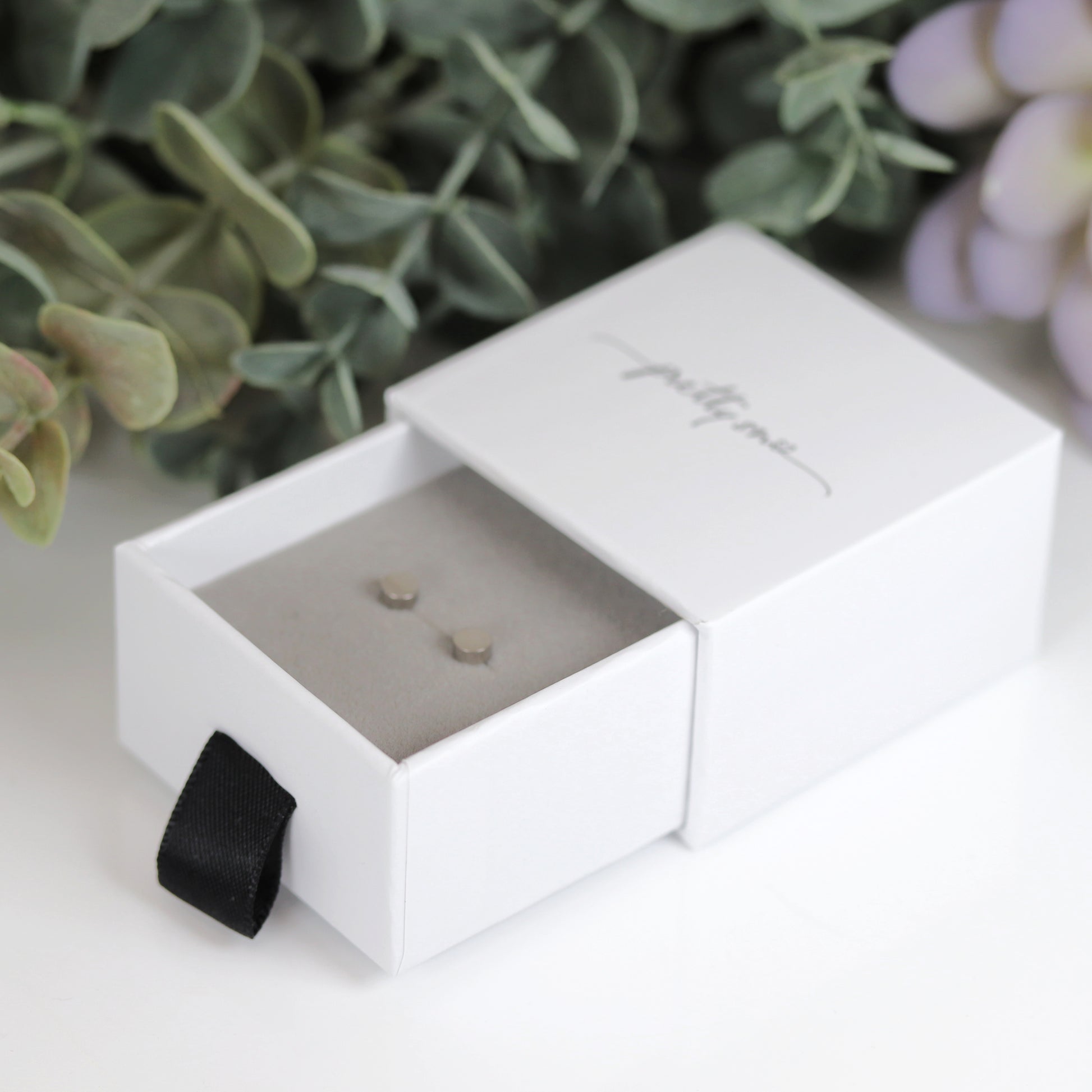 A pair of round dot stud earrings in silver and gold colors, placed on a white surface with foliage in the background.