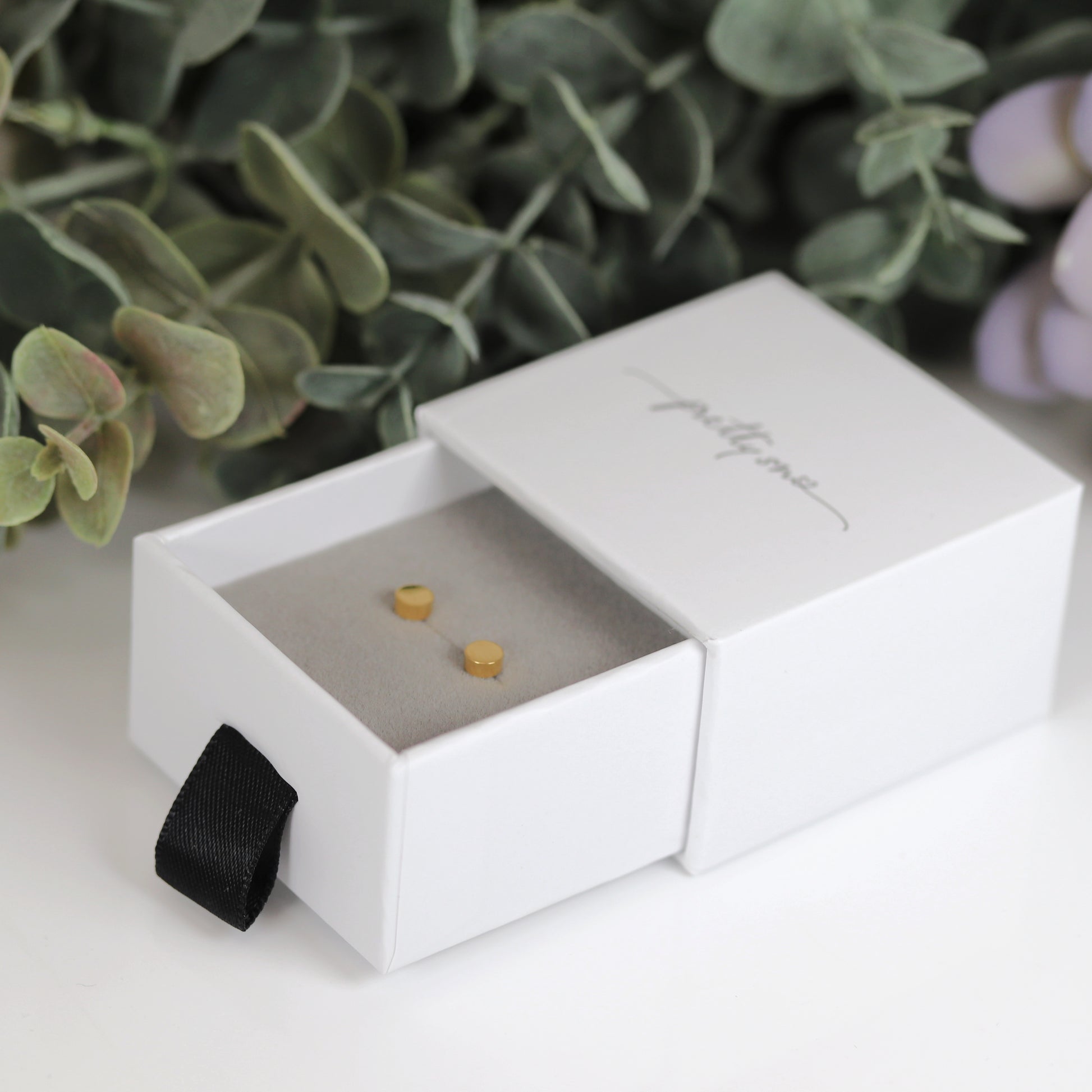 A pair of round dot stud earrings in silver and gold colors, placed on a white surface with foliage in the background.